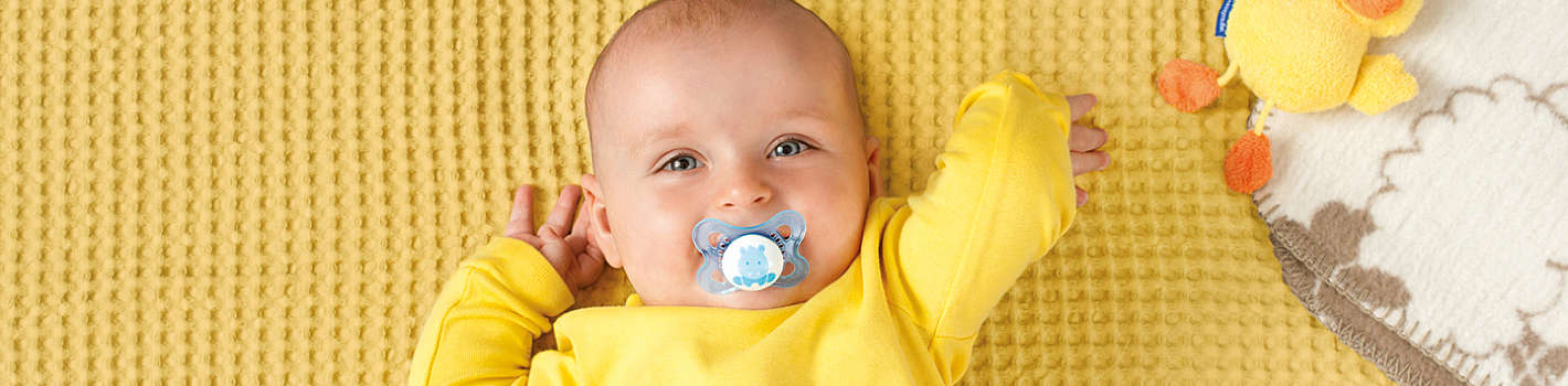 Fröhliches Baby liegt auf gelber Decke am Rücken und hat hellblauen MAM Original Schnuller mit Tiermotiv im Mund