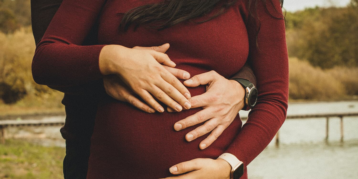 De futurs parents se tiennent en extérieur, les mains des deux partenaires posées sur le ventre de la future maman. 
