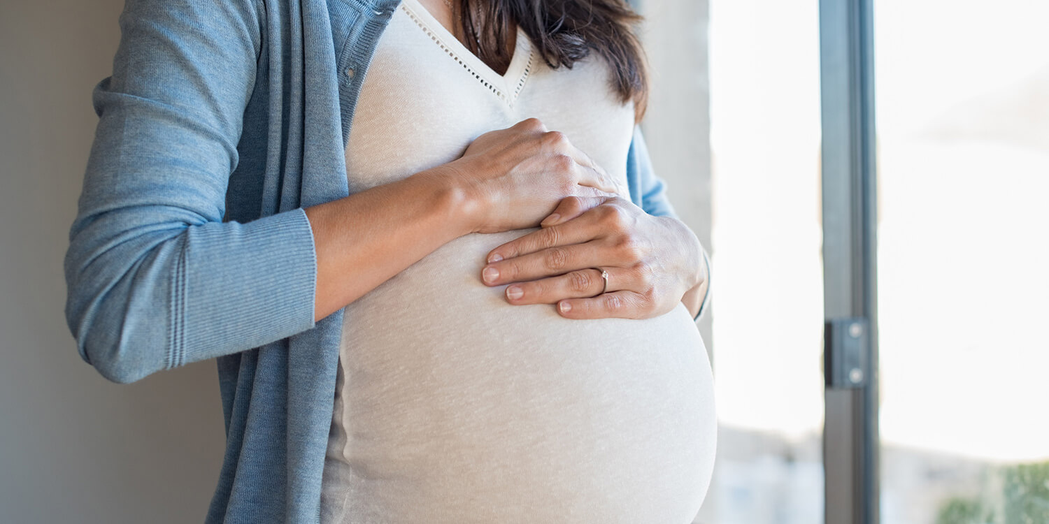 Gravid kvinna lägger händerna på sin gravidmage och tittar ut genom fönstret