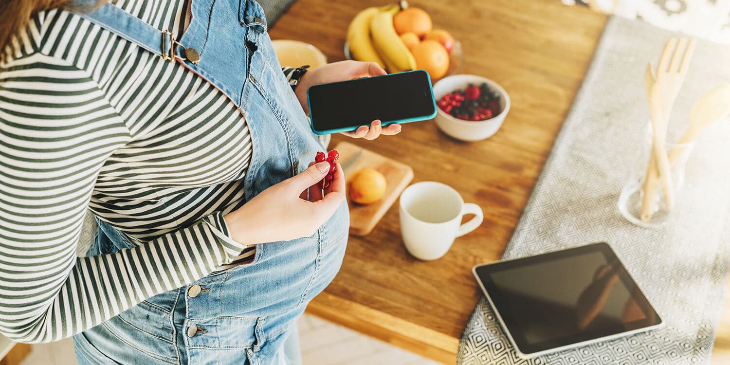 Pregnant woman holds her mobile phone in one hand and fruit in the other, a dining table with fruit is in the background.