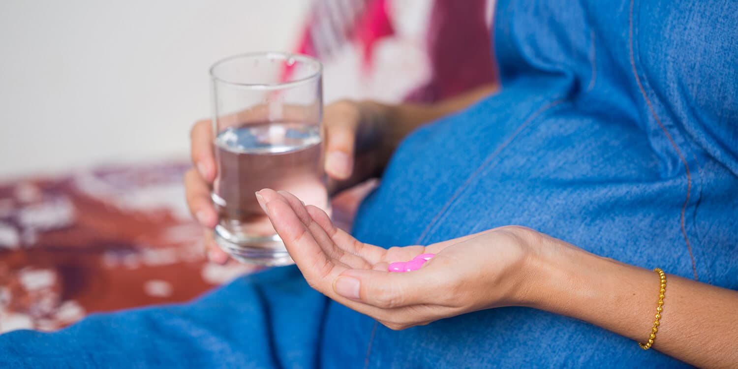 Schwangere Frau sitzt auf einer Couch und hält Tabletten und ein Glas Wasser in ihren Händen. 