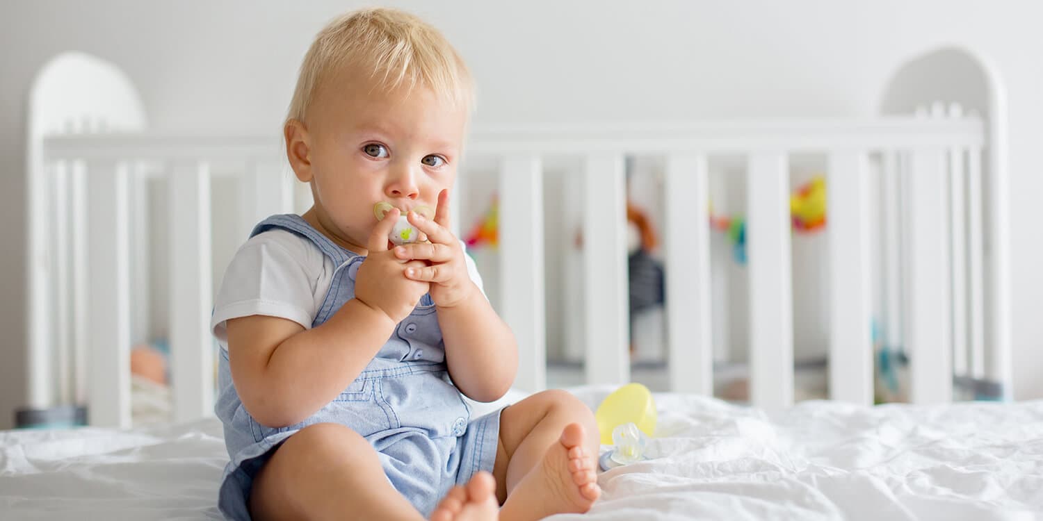 Toddler sits on bed and has pacifier in its mouth, it grasps the pacifier with both hands
