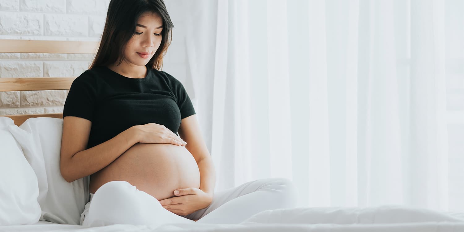 Gravid kvinna sitter på sängen och håller försiktigt om sin gravidmage.