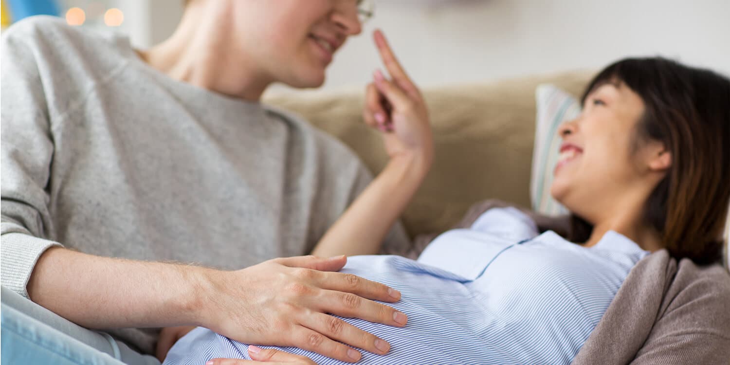 Um casal grávido aconchega-se no sofá e ri; o homem coloca as mãos sobre a barriga de grávida da mulher