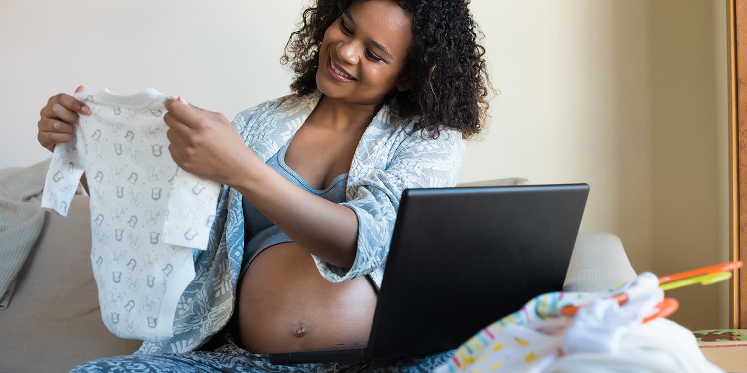 Woman sitting on the couch, shopping online for her baby while she is holding up and looking at a babyromper.