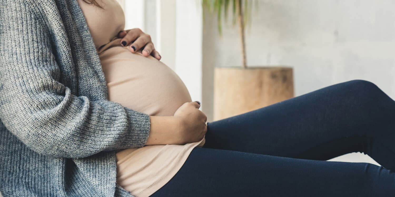 Uma mulher sentada sobre uma manta no chão coloca as mãos sobre a sua barriga de grávida.